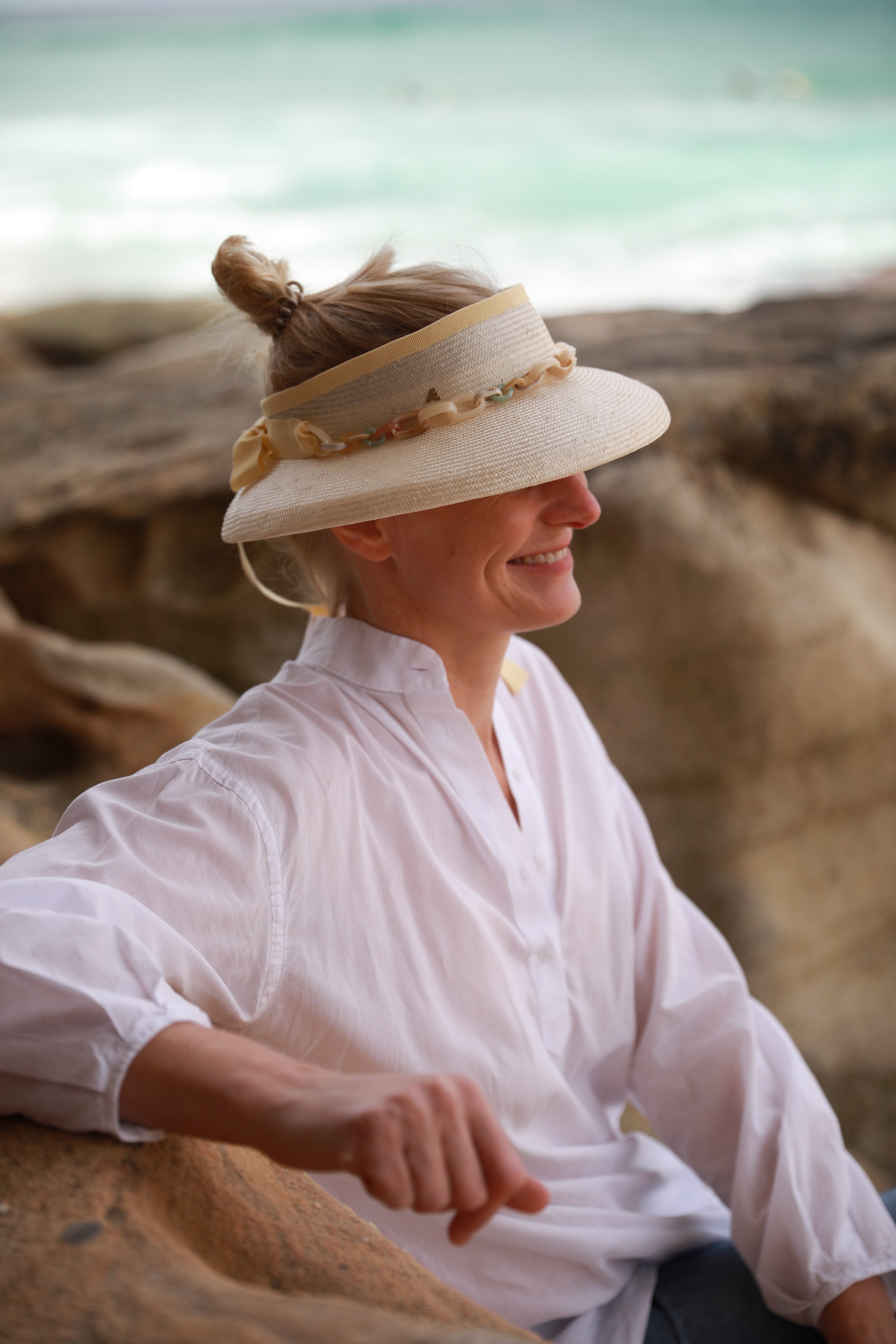 A woman wearing a white visor hat with a turned-down brim and a multicolored chain around the head, sitting by a rocky beach with the ocean in the background.
