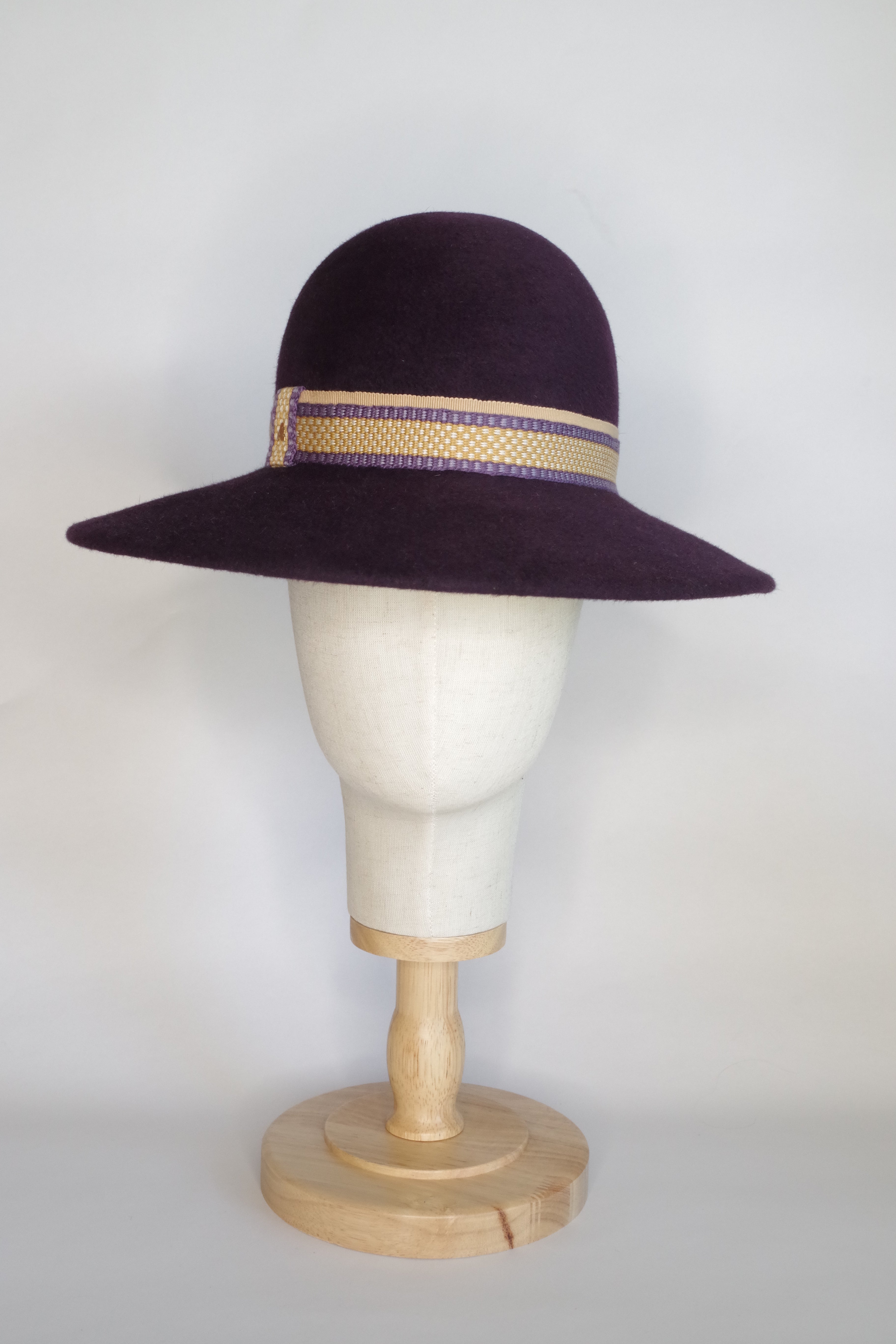 A deep purple velour fur felt hat with a handwoven ribbon in warm yellow and purple displayed on a mannequin head.