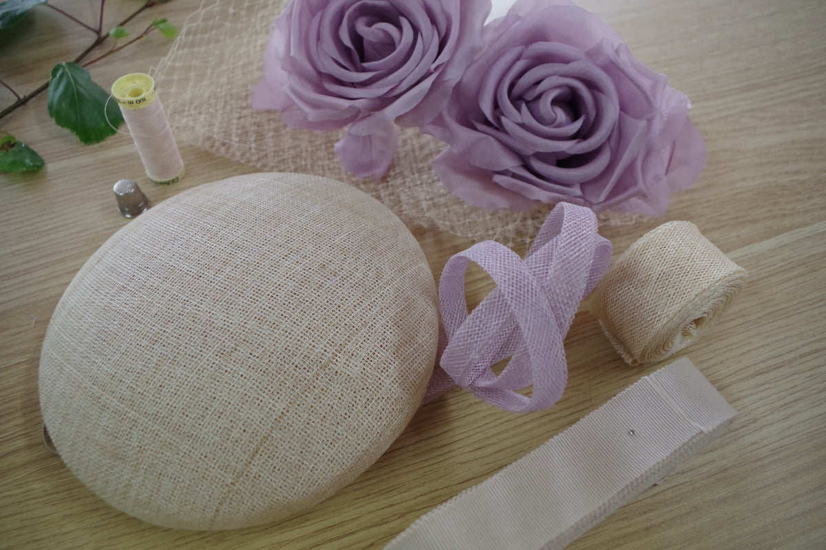 Millinery materials laid out on a wooden table in the MICHAETT studio in Bowral – a sinamay hat base, lilac roses, veiling, thread and rolls of sinamay and petersham ribbon, ready for a workshop.